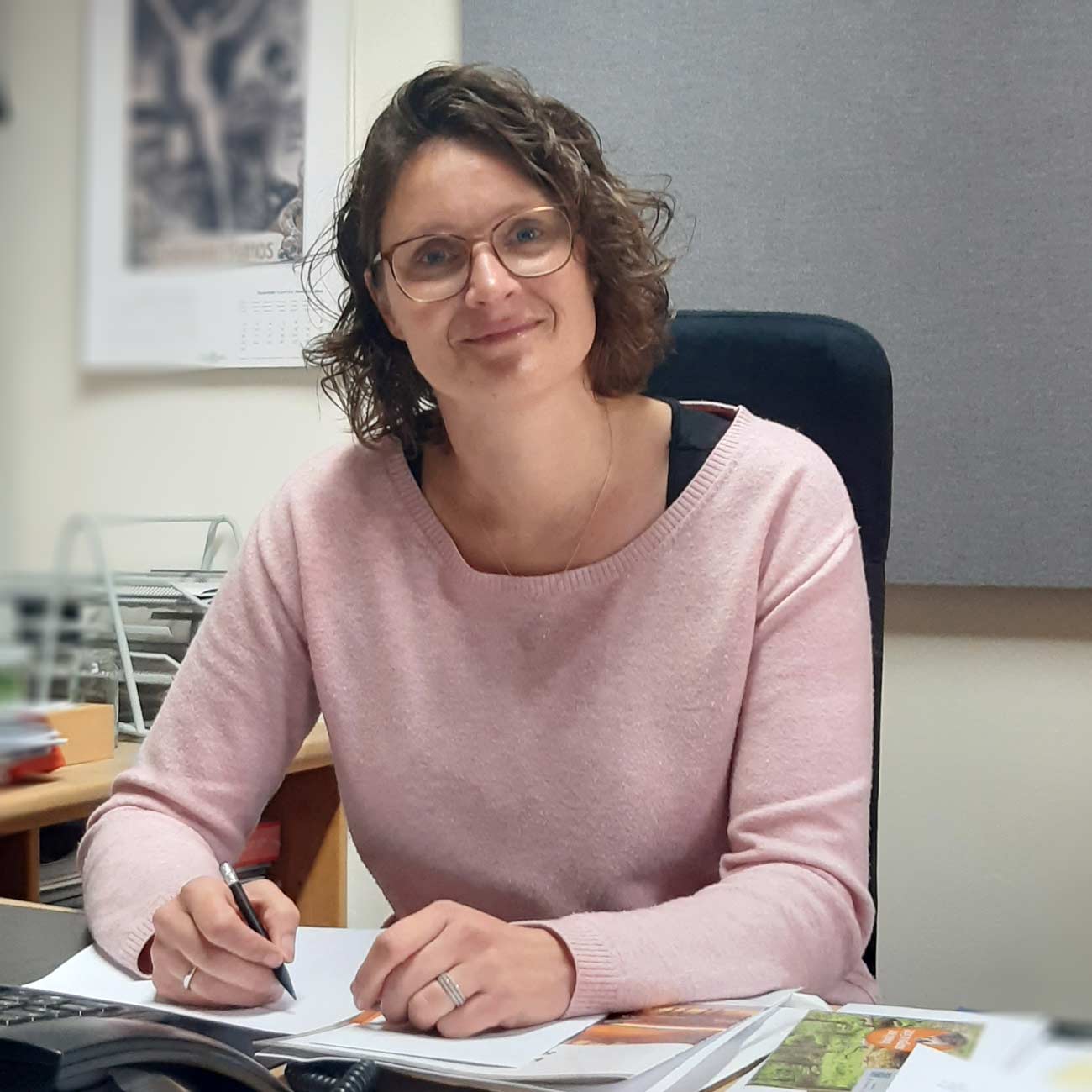 Frau mit Brille sitzt lächelnd am Schreibtisch und schreibt auf Papier in einem Büro mit Kalender im Hintergrund​​​​​​​​​​​​​​​​​​​​​​​​​​​​​​​​​