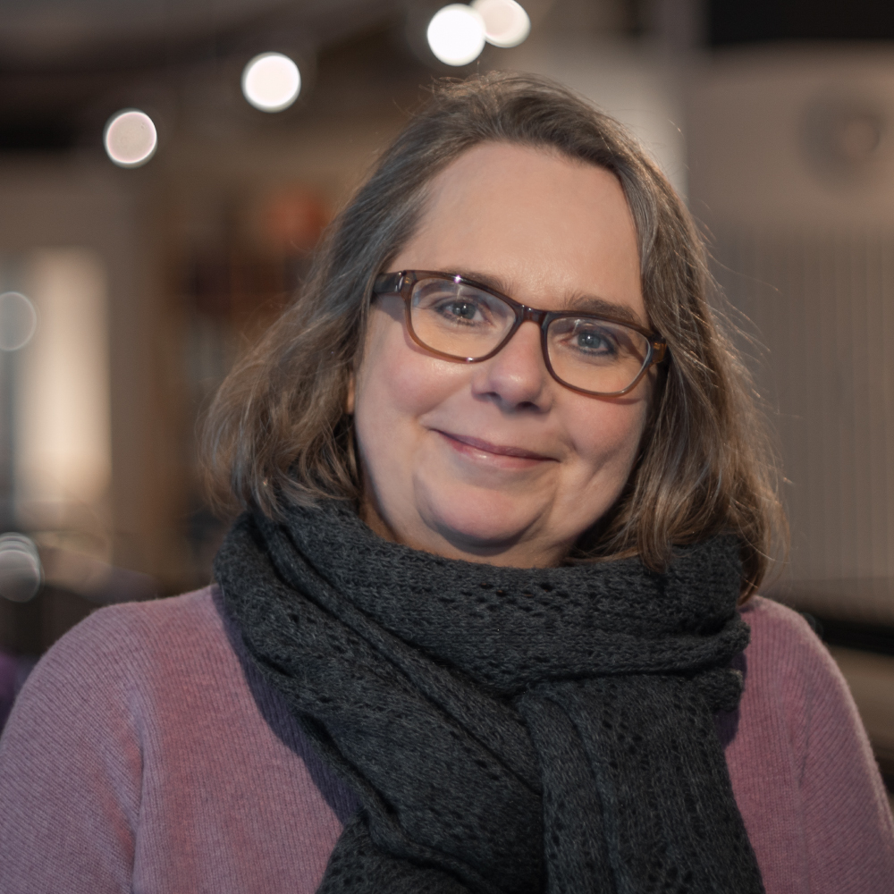 Nahaufnahme einer lächelnden Frau mit Brille, grauem Haar und schwarzem Schal in gemütlicher Innenraumumgebung​​​​​​​​​​​​​​​​​​​​​​​​​​​​​​​​​​​